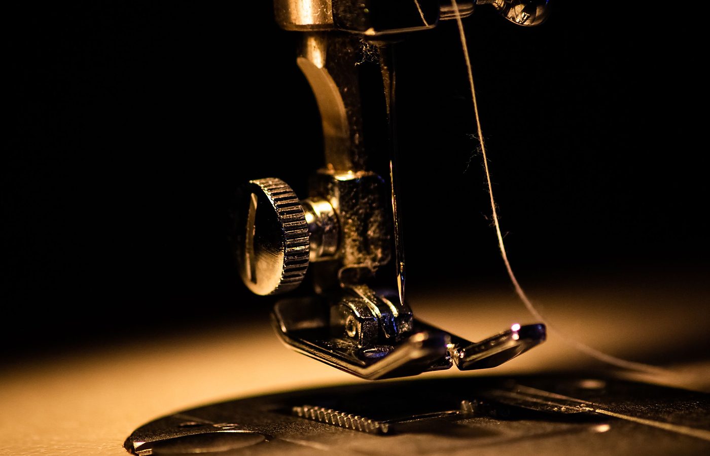 Licensed stock photograph: the brass presser foot of a sewing machine under a warm lamp, with a threaded needle above the plate.